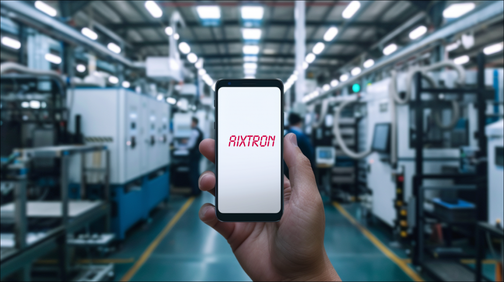 eine Hand, die ein Smartphone mit dem Schriftzug AIXTRON auf dem Bildschirm hält, im Hintergrund ist eine unscharfe Fabrikhalle mit industriellen Maschinen 