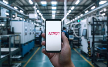 eine Hand, die ein Smartphone mit dem Schriftzug AIXTRON auf dem Bildschirm hält, im Hintergrund ist eine unscharfe Fabrikhalle mit industriellen Maschinen 