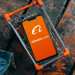 Ein Smartphone mit dem Bildschirm, der das Alibaba-Logo zeigt, liegt in einem Miniatur-Einkaufswagen 