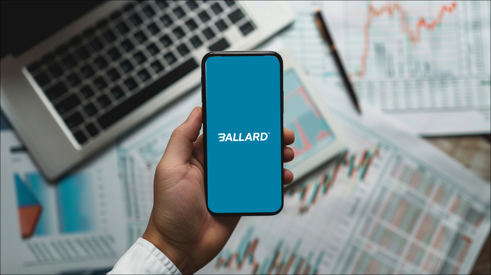 Eine Hand hält ein Smartphone mit dem Logo von Ballard Power Systems auf dem Display vor einem Hintergrund mit Finanzdiagrammen und einem Laptop.