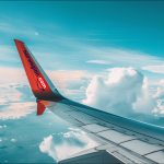 die Flügelspitze eines Flugzeugs mit dem Schriftzug norwegian.com und einem markanten roten Endteil, das gegen einen klaren Himmel und weiße Wolken abgehoben ist.