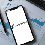 Ein Arbeitsplatz mit einem Smartphone, auf dessen Display das Logo von RHEINMETALL, neben Papieren mit Diagrammen