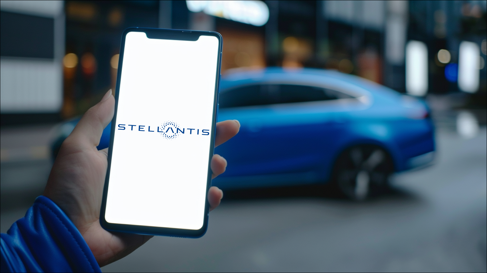 Eine Hand hält ein Smartphone mit eingeschaltetem Bildschirm, der das Logo von Stellantis zeigt, vor einem unscharfen Hintergrund mit einem blauen Auto.