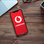 Ein Smartphone mit dem Vodafone-Logo auf dem Bildschirm liegt neben einem Laptop, einer Kamera und anderen Bürogegenständen auf einem Holztisch.
