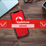 Ein Smartphone mit dem Vodafone-Logo auf dem Bildschirm liegt neben einem Laptop, einer Kamera und anderen Bürogegenständen auf einem Holztisch, mit einem Vodafone NEWS Banner 