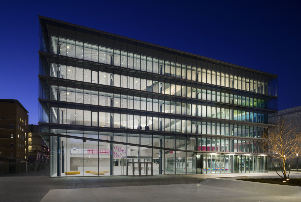 Ein modernes, mehrstöckiges Bürogebäude mit vollständig verglaster Fassade, das bei Nacht hell erleuchtet ist. Das Gebäude wirkt offen und transparent, mit deutlich sichtbaren Etagen und Innenräumen. Vor dem Eingang stehen Sitzgelegenheiten und ein kahler Baum, der von einer Bodenleuchte angestrahlt wird. Der Himmel ist tiefblau und klar.