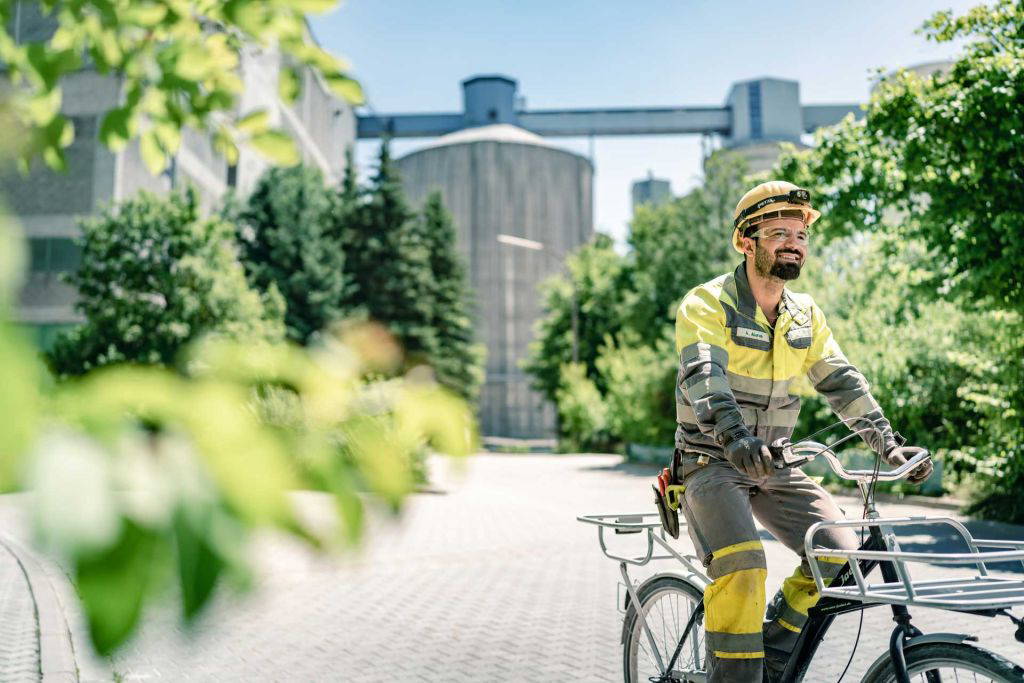 Ein Mann in gelber Arbeitskleidung und Helm fährt mit einem Lastenfahrrad durch ein grünes, sonniges Industriegelände. Im Hintergrund sind Silos und Fabrikgebäude zu sehen.