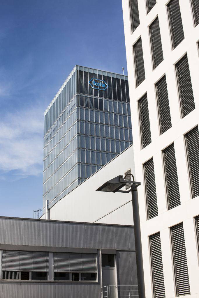 Blick auf den modernen Roche-Turm mit Logo in Basel bei blauem Himmel, teilweise verdeckt durch angrenzende Bürogebäude.