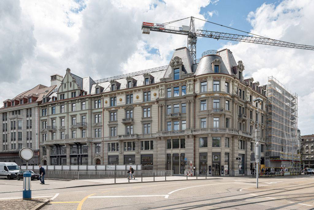 Straßenansicht eines renovierten historischen Gebäudes mit Fassadendetails, großen Fenstern und Dachgauben am Bahnhofplatz in Zürich. Im Hintergrund ragt ein Baukran über das Gebäude, rechts ist ein eingerüsteter Gebäudeteil zu sehen. Einige Passanten und Fahrzeuge sind auf der Straße unterwegs.