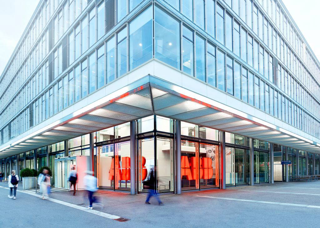 Außenansicht des modernen ABB-Hauptgebäudes in Zürich mit markanter Glasfassade. Das ABB-Logo ist mehrfach hinter den Eingangsbereichen zu sehen. Zahlreiche Passanten bewegen sich vor dem Gebäude – es herrscht geschäftiges Treiben.