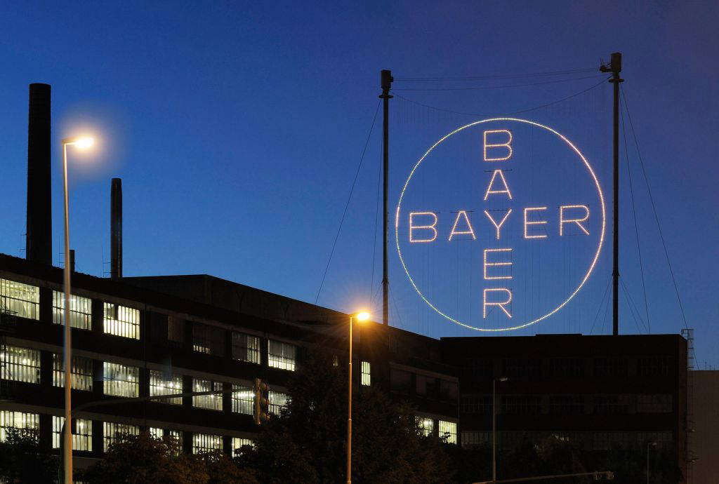Das Foto zeigt das beleuchtete Bayer-Kreuz über einem Werksgebäude in Leverkusen bei Nacht. Die ikonische Leuchtreklame mit dem Schriftzug „BAYER“ in vertikaler und horizontaler Ausrichtung ist über Seilen zwischen zwei Masten gespannt. Im Vordergrund sind Fabrikgebäude mit erleuchteten Fenstern und mehrere Straßenlaternen zu sehen. Der Himmel ist tiefblau und wolkenlos.