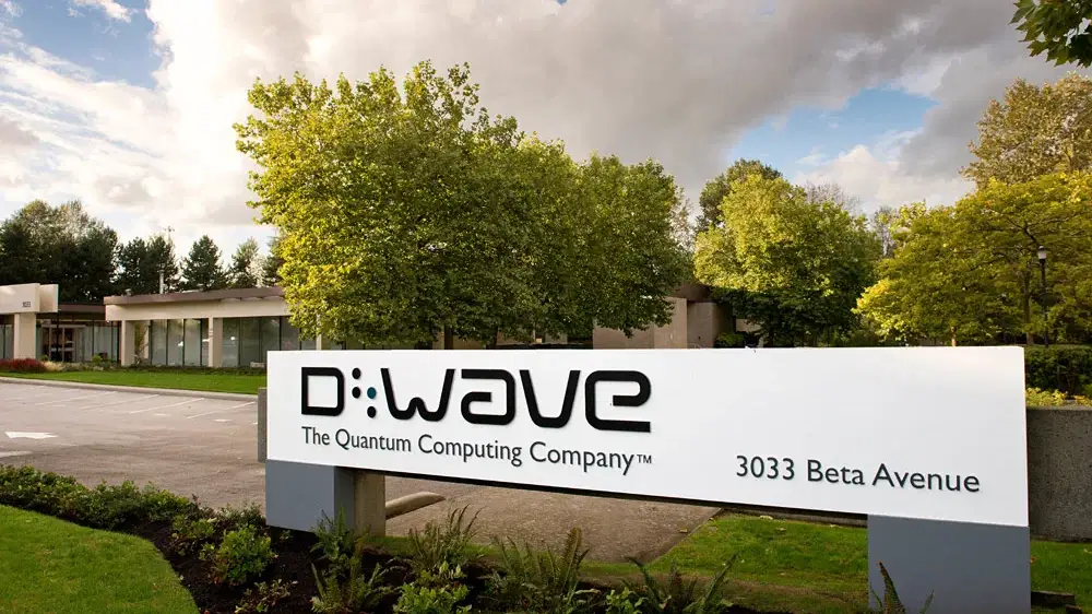 Firmensitz von D-Wave mit weißem Eingangslogo „The Quantum Computing Company“ auf grün bepflanztem Gelände bei bewölktem Himmel.