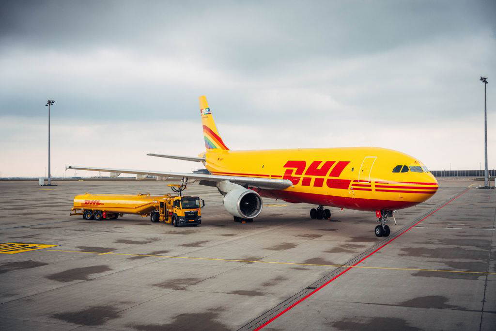 Ein DHL-Frachtflugzeug wird auf einem Flughafenrollfeld betankt. Im Vordergrund steht ein gelber Tankwagen mit DHL-Logo, der das Flugzeug versorgt. Die Szene spielt sich an einem wolkigen Tag auf einem weitläufigen Flughafenareal ab – Symbolbild für globale Logistik und Frachttransport.