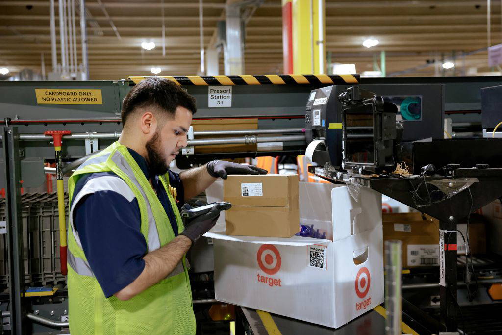 Mitarbeiter eines Target-Logistikzentrums scannt ein Paket an einer Versandstation. Er trägt eine Sicherheitsweste und Handschuhe. Im Hintergrund ist ein automatisiertes Fördersystem sichtbar. Ein großer Target-Karton steht bereit zur Befüllung.