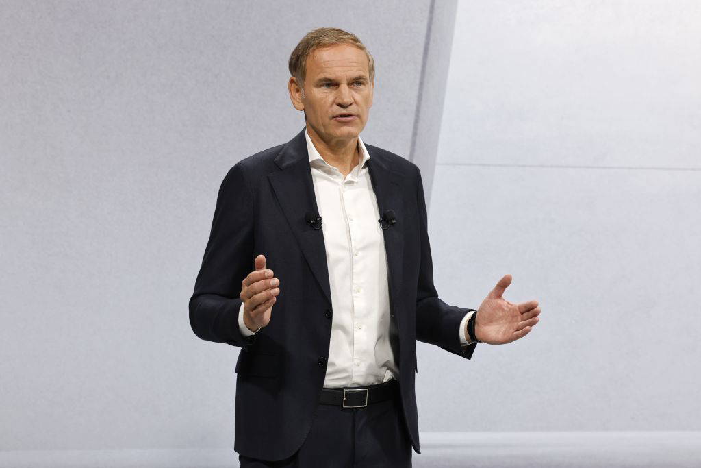 Oliver Blume, Vorstandsvorsitzender der Porsche AG, spricht auf einer Bühne. Er trägt einen dunklen Anzug und ein weißes Hemd, gestikuliert mit beiden Händen und trägt zwei Ansteckmikrofone. Der Hintergrund ist schlicht und hell.