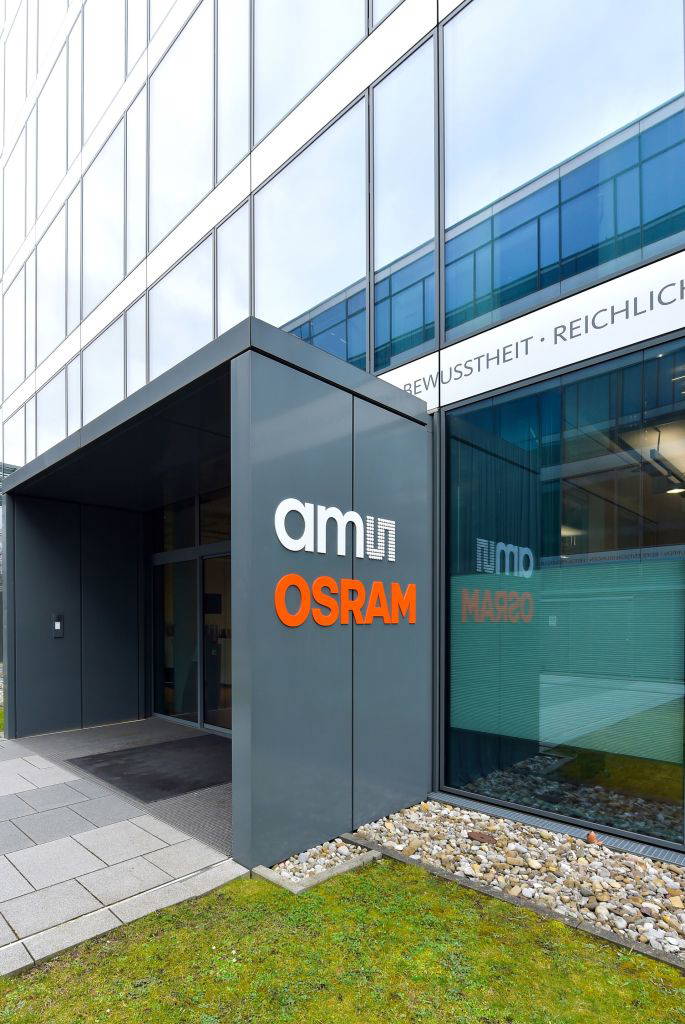 Gebäudeeingang mit Fokus auf das „ams OSRAM“-Logo und einem Blick auf die gepflegte Außenanlage mit Kiesbeeten und Rasenfläche. Im Fenster spiegeln sich Gebäudeteile und Schriftzüge.