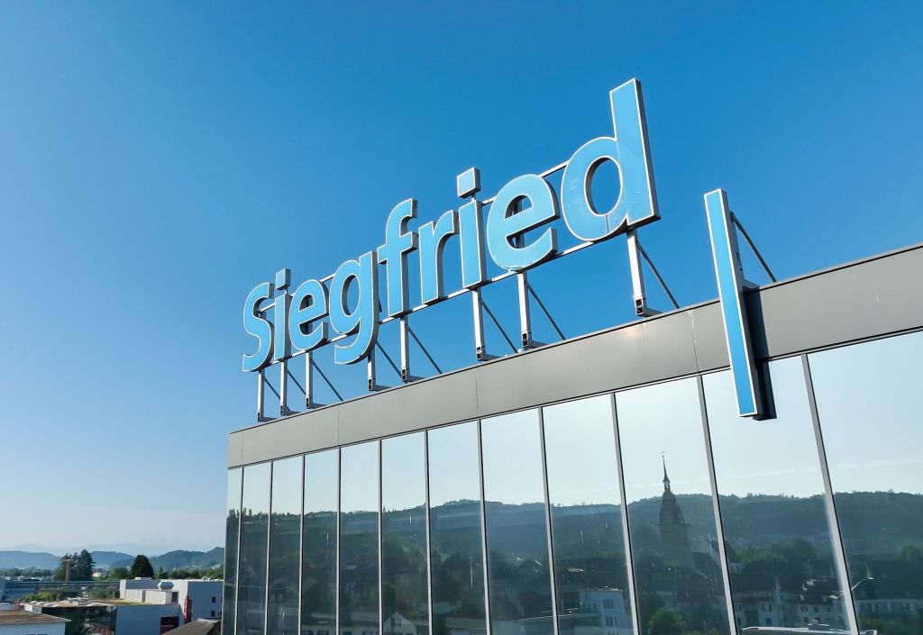 Ein modernes Firmengebäude mit reflektierender Glasfassade, auf dessen Dach in großen hellblauen Buchstaben der Schriftzug „Siegfried“ angebracht ist; im Hintergrund ist eine hügelige Landschaft mit vereinzelten Gebäuden und einem Kirchturm zu sehen, der sich in der Glasfassade spiegelt. Der Himmel ist klar und blau.