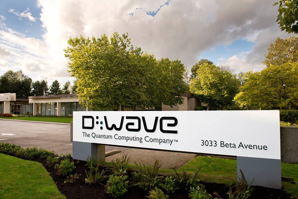 Firmensitz von D-Wave mit weißem Eingangslogo „The Quantum Computing Company“ auf grün bepflanztem Gelände bei bewölktem Himmel.