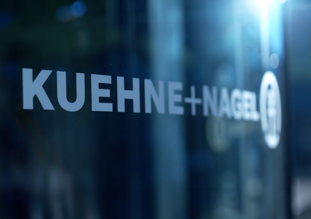 Glasfassade mit dem weiß aufgedruckten Firmenlogo „KUEHNE+NAGEL“ in Großbuchstaben; rechts daneben das runde Unternehmenssymbol. Die Spiegelung im Glas zeigt unscharfe Umrisse eines modernen Gebäudes bei Tageslicht.