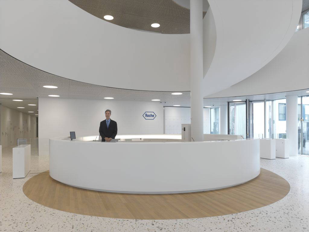 Empfangsbereich von Roche mit rundem weißen Tresen, einem Mitarbeiter im Anzug und dem Roche-Logo an der Wand.
