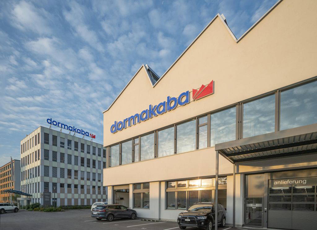 Außenansicht der Dormakaba-Zentrale mit Firmenlogo auf zwei Gebäuden und Parkplatz im Vordergrund.