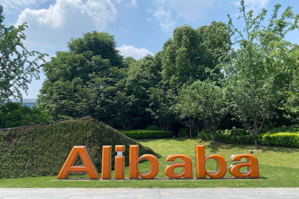 Ein großes, orangefarbenes Alibaba-Logo steht auf einer gepflegten Rasenfläche vor einer mit Efeu bewachsenen Wand. Im Hintergrund sind zahlreiche grüne Bäume und ein blauer Himmel mit einigen Wolken zu sehen. Die Szene vermittelt eine ruhige, naturverbundene Unternehmensumgebung.