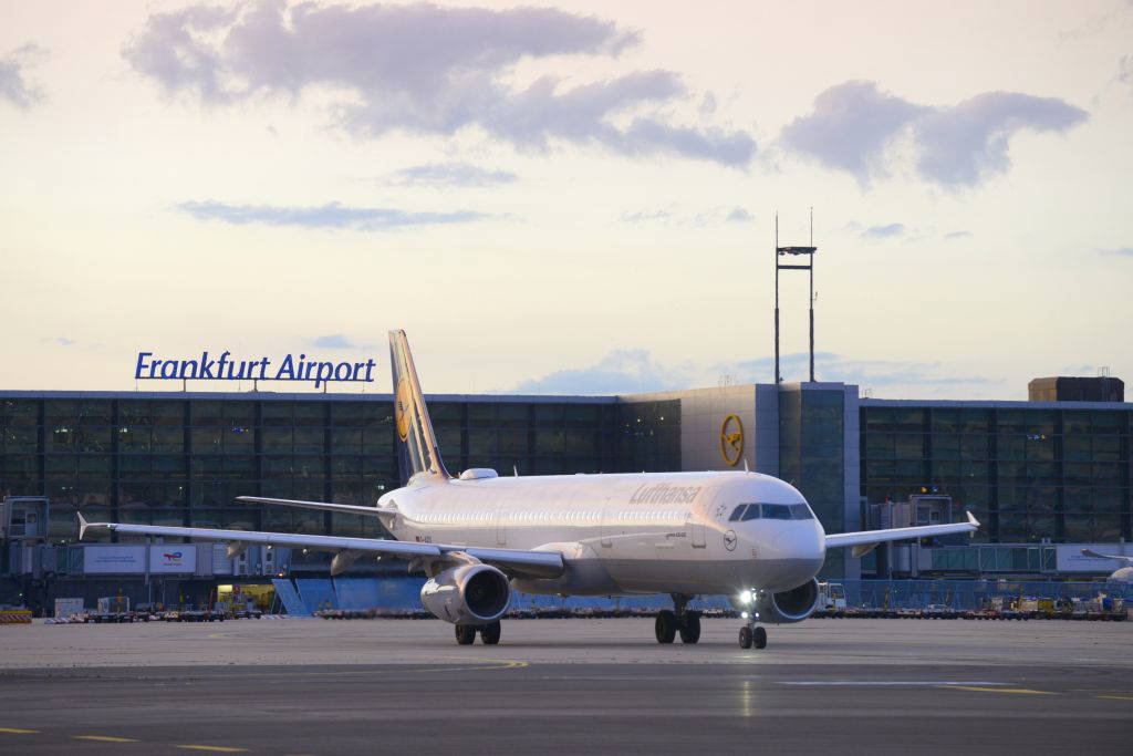 Ein Lufthansa-Flugzeug rollt bei Sonnenuntergang über das Vorfeld des Flughafens Frankfurt. Im Hintergrund ist das Terminal mit dem Schriftzug „Frankfurt Airport“ gut sichtbar. Der Himmel zeigt sanfte Wolken vor einem abendlichen Licht. Die Szene vermittelt einen ruhigen Moment im internationalen Flugverkehr. © Fraport AG