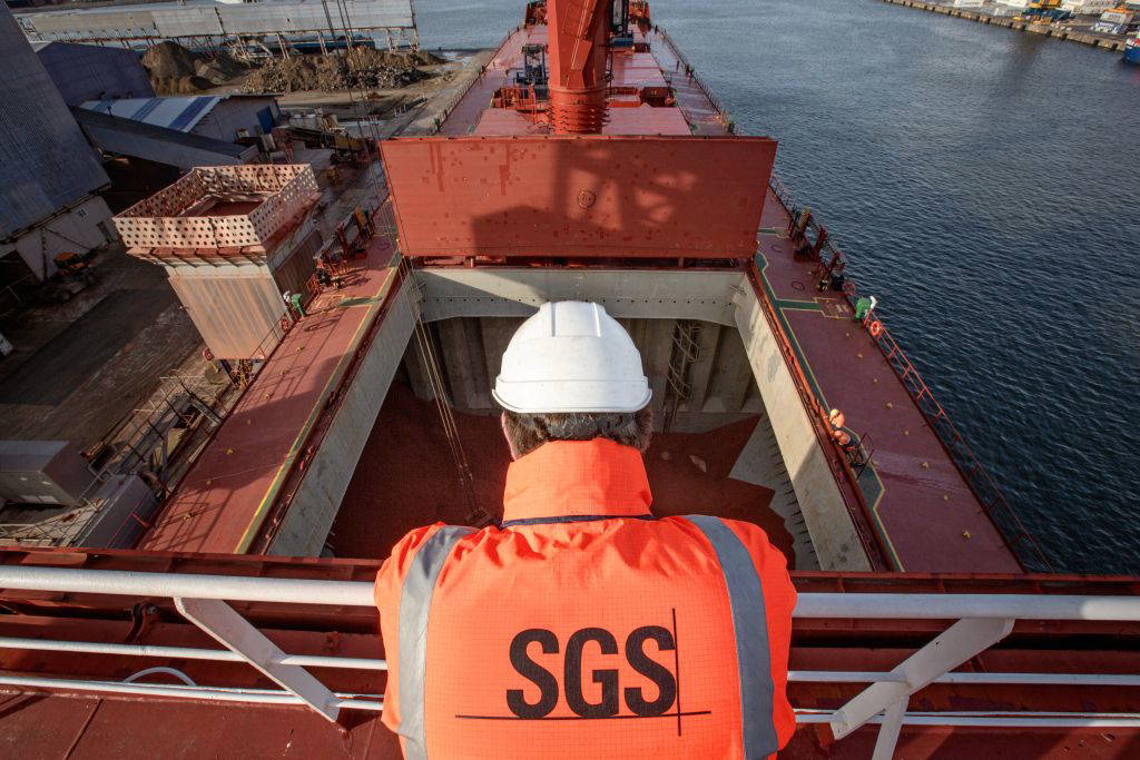 Eine Person mit Helm und oranger Arbeitsjacke mit „SGS“-Logo blickt von einer erhöhten Plattform auf ein großes Frachtschiff im Hafen. Das Schiff liegt in einem industriellen Hafengebiet, und das Bild zeigt den Laderaum, verschiedene Kräne und ein Hafenbecken im Hintergrund. © SGS SA