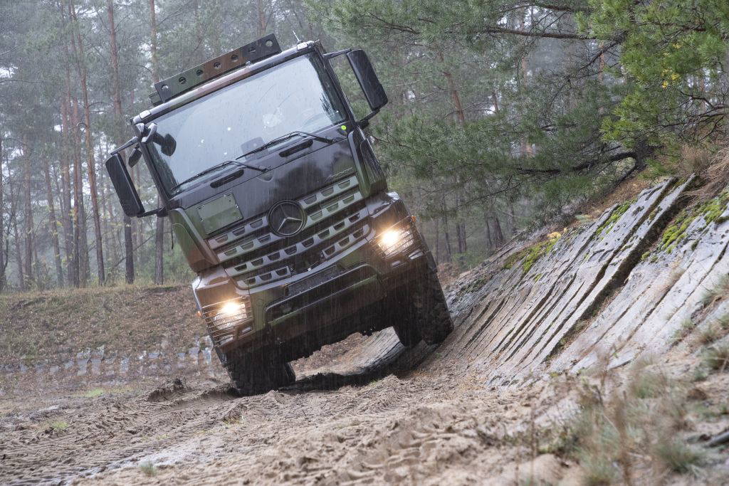 Ein militärisch ausgerüsteter Mercedes-Benz Arocs 6x6 Lkw fährt durch einen matschigen, unwegsamen Waldweg. Das Fahrzeug, in Tarnfarben lackiert, zeigt seine Geländetauglichkeit bei Nässe und starkem Regen. Die Aufnahme zeigt den Lkw steil geneigt an einem Hang, frontal in schwierigen Geländepassagen © Daimler Truck Holding AG