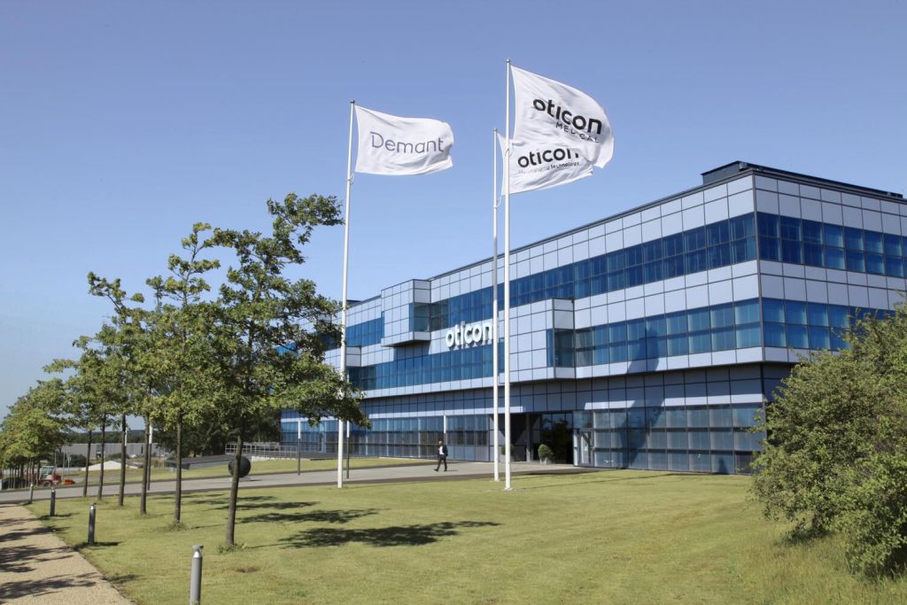Moderne Firmenzentrale von Oticon, einer Marke der Demant-Gruppe, mit gläserner Fassade und drei wehenden Fahnen mit den Logos von Demant und Oticon. Im Vordergrund eine gepflegte Rasenfläche mit Bäumen. © Demant A/S