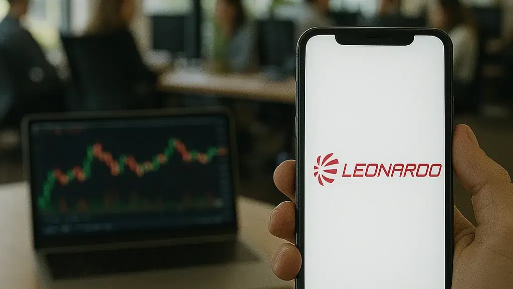 In einem modernen, lichtdurchfluteten Großraumbüro hält eine Hand ein Smartphone mit dem rot leuchtenden Leonardo-Logo auf dem Bildschirm. Im Hintergrund arbeitet eine Gruppe von Menschen an Schreibtischen, während auf einem Laptop ein Aktienchart angezeigt wird.