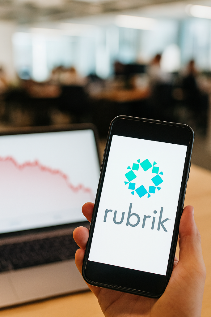 Eine Person hält ein Smartphone mit dem Rubrik-Logo im Display. Im Hintergrund sieht man einen Laptop, der einen fallenden Aktienchart ohne Beschriftung zeigt. Die Szene spielt sich in einem geschäftigen Großraumbüro ab.