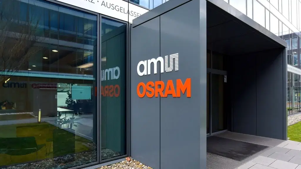Gebäudeeingang mit Fokus auf das „ams OSRAM“-Logo und einem Blick auf die gepflegte Außenanlage mit Kiesbeeten und Rasenfläche. Im Fenster spiegeln sich Gebäudeteile und Schriftzüge.