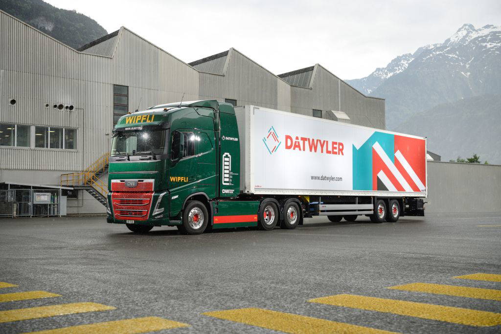 Ein großer LKW der Firma Wipfli, beladen mit einem Container mit dem Logo und Schriftzug von Dätwyler, steht vor einem grauen Industriegebäude. Im Hintergrund sind Berge mit Schnee auf den Gipfeln zu sehen. Der Himmel ist bewölkt und der Boden nass – es hat offenbar gerade geregnet. © Dätwyler Holding AG