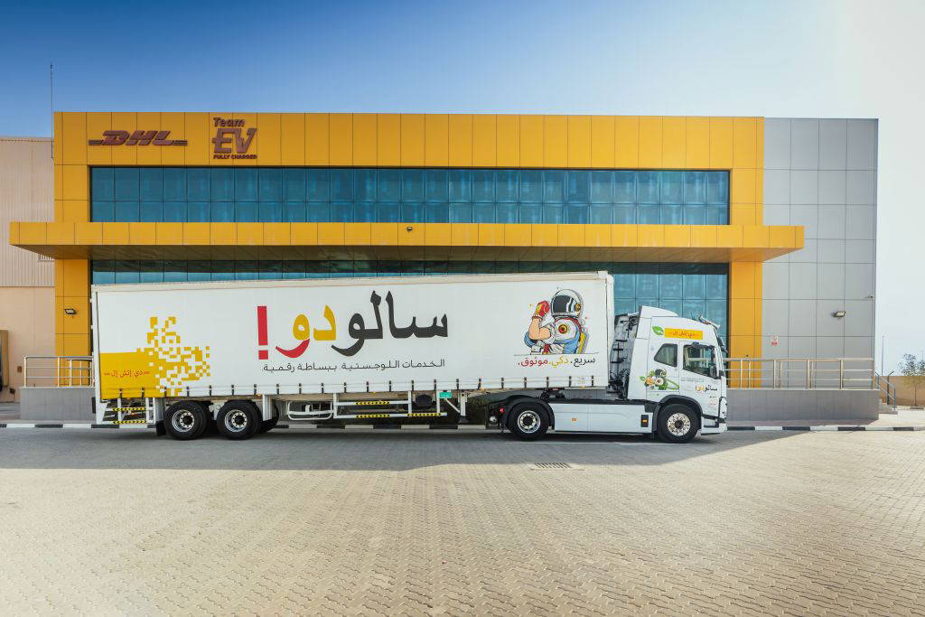 Ein moderner DHL-LKW mit arabischer Beschriftung und einer Comic-Zeichnung eines Astronauten auf der Seite steht vor einem großen gelb-grauen Logistikzentrum. Der Himmel ist klar und blau, der Boden mit Pflastersteinen bedeckt. Die Szene wirkt sauber, modern und professionell. © DHL Group AG