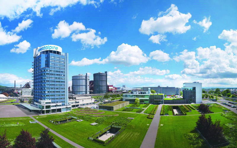 Blick auf den Hauptsitz von Voestalpine in Linz: Das markante Hochhaus mit Logo ist umgeben von einer weitläufigen grünen Parkanlage. Im Hintergrund sind Industrieanlagen und weitere Firmengebäude zu sehen, während der Himmel von weißen Wolken durchzogen ist.