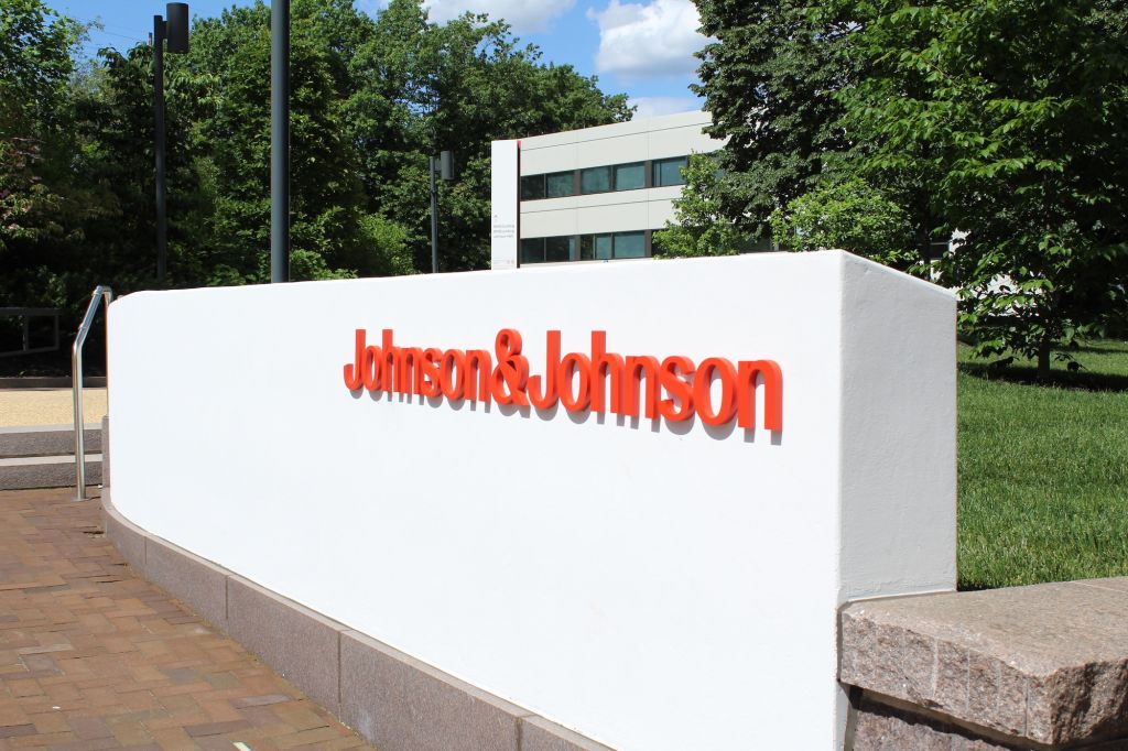 Johnson & Johnson-Aktie: Gleich zwei Erfolge! Außenansicht des Johnson & Johnson-Firmenschilds an einer weißen Mauer mit rotem Firmenlogo, im Hintergrund grüne Bäume und Bürogebäude.
