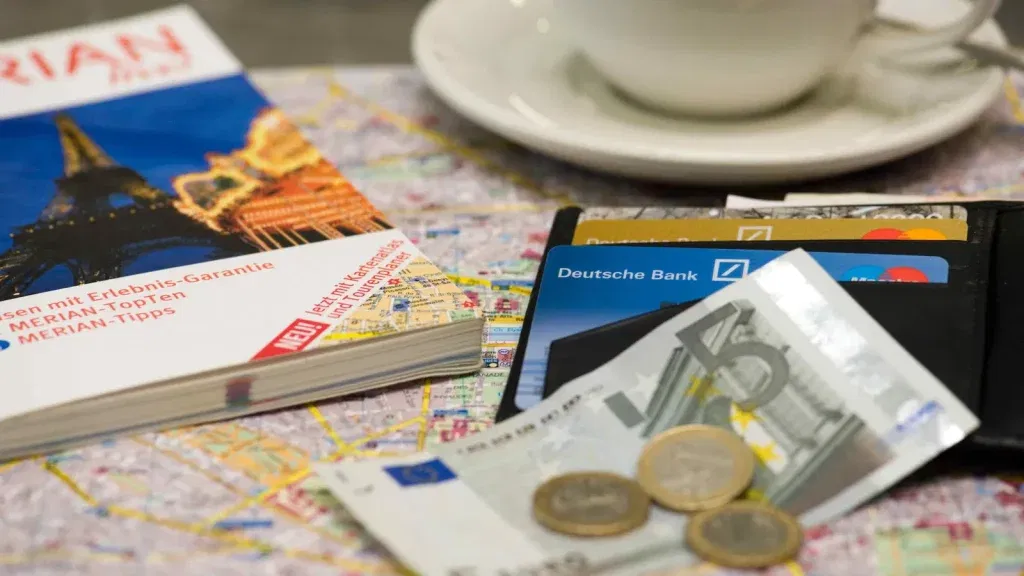 Reiseführer, Geldbörse mit Deutsche-Bank-Karten und Euro-Bargeld liegen auf einem Stadtplan neben einer Kaffeetasse – Symbol für Reiseplanung und Finanzen.